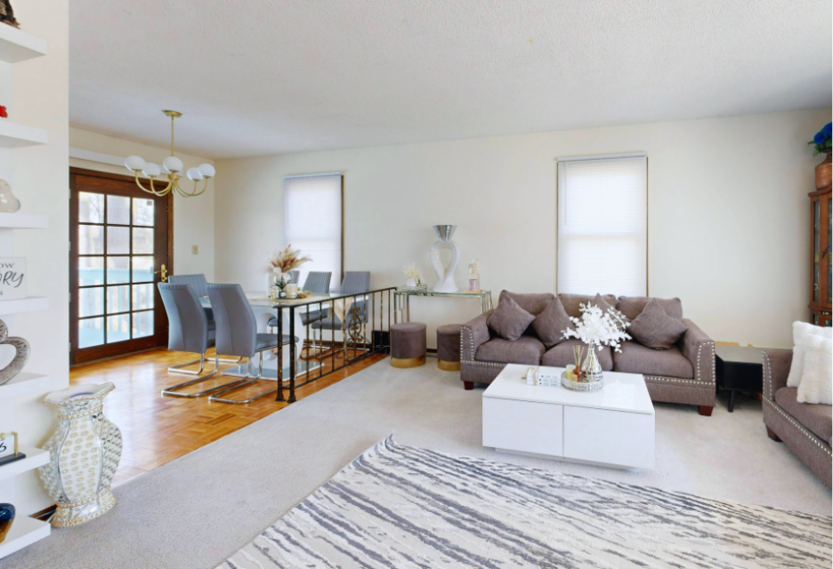 Bright living room with a brown sofa, white coffee table, patterned area rug, dining table and chairs, and decorative shelving.